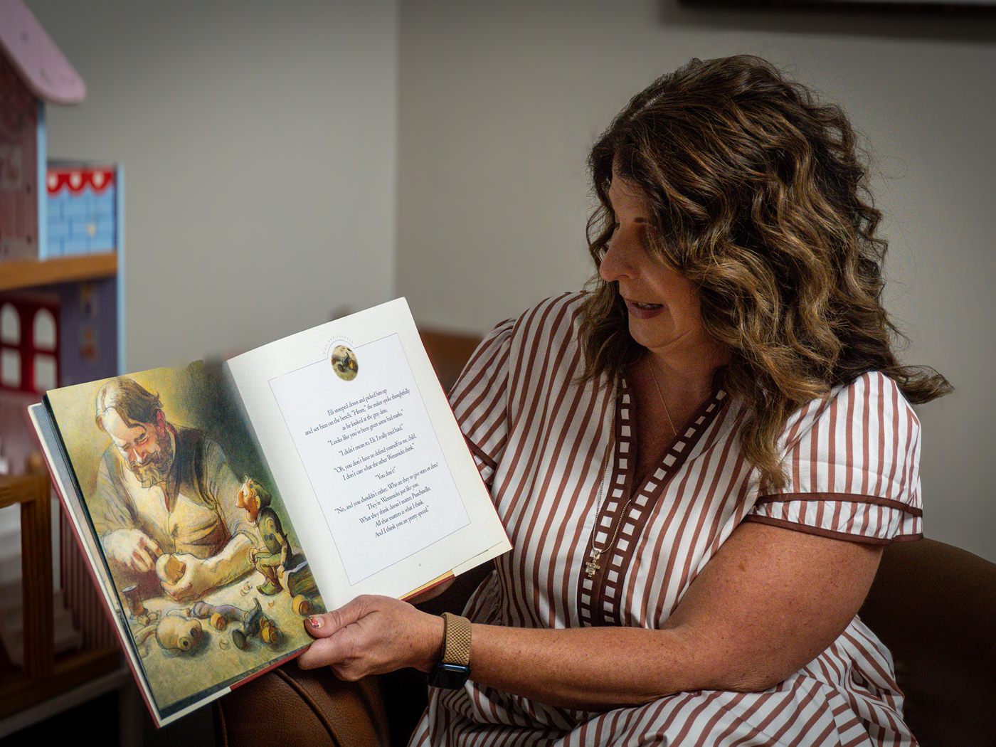 DeeDee seated in her counseling room, reading aloud from an open illustrated picture book, with a doll house on the shelf behind her.