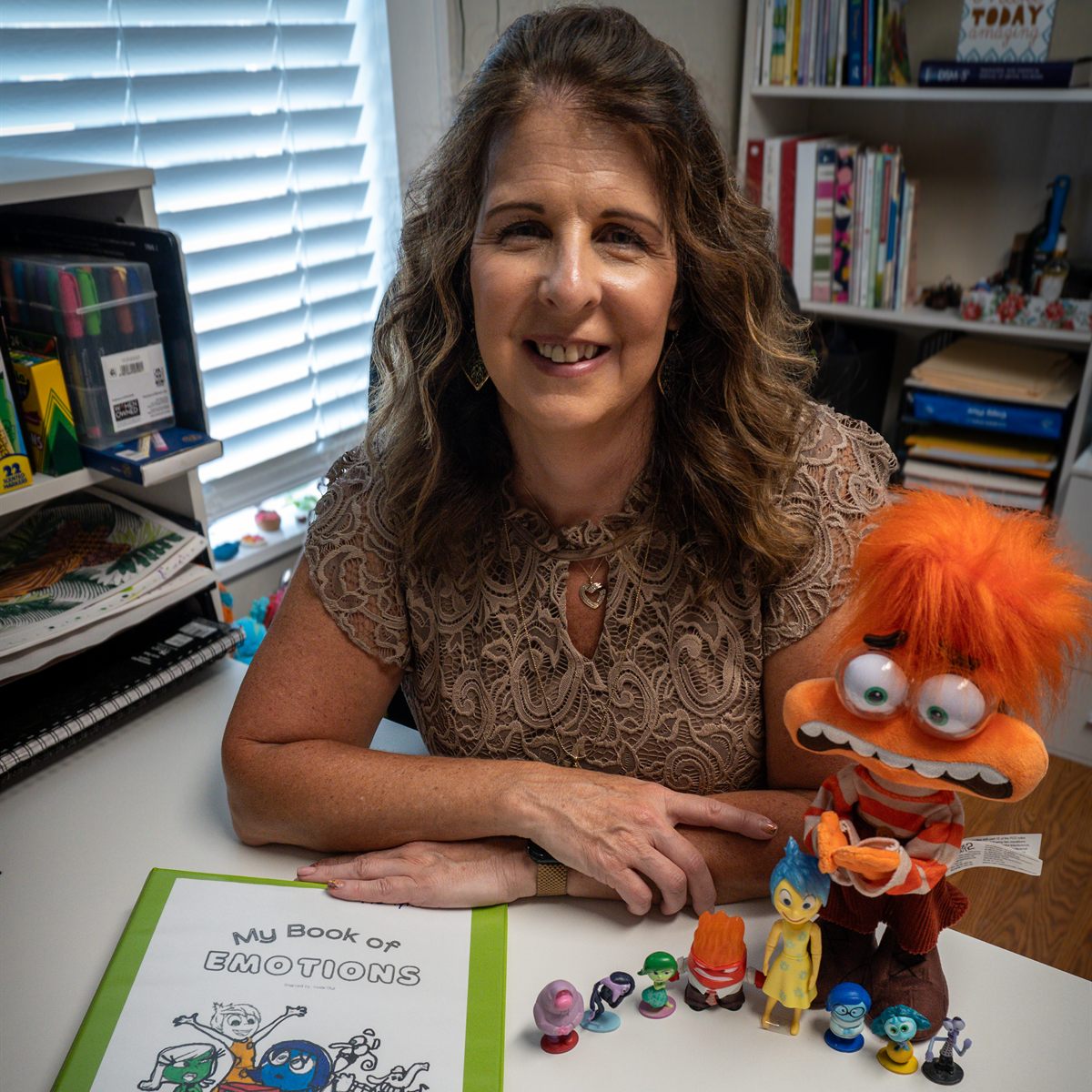 DeeDee smiling at her office desk beside an orange puppet, a binder titled My Book of Emotions, and a row of feelings figurines she uses in play therapy.