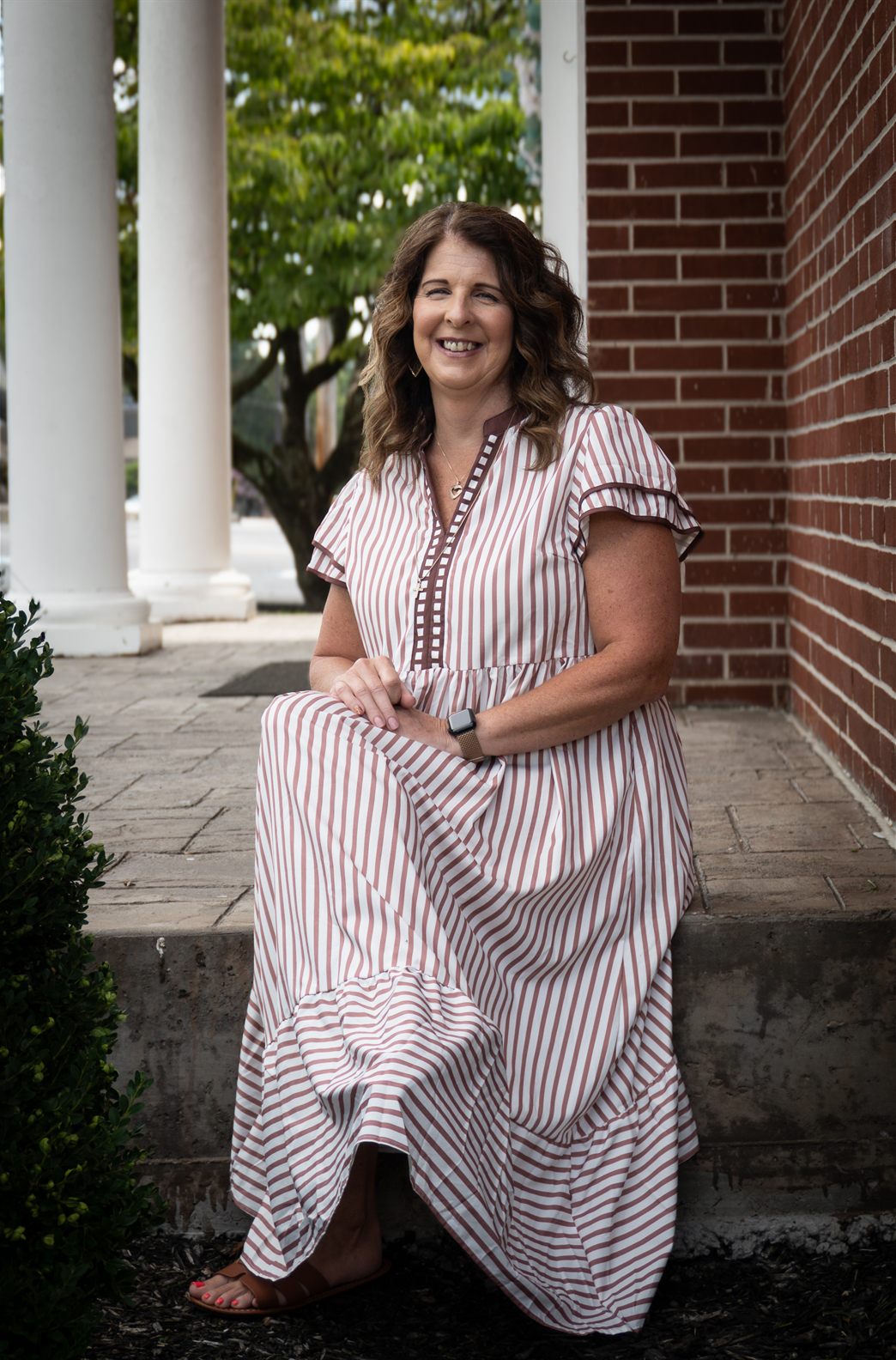 DeeDee Miller seated on a step outside a brick building with white porch columns, smiling.